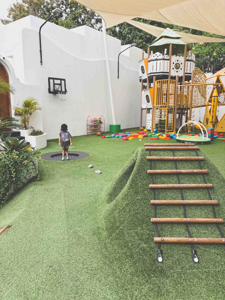 Pangolin Kids Club outside playground. A child stands near a trampoline and basketball hoop in a playground with artificial grass, a small climbing hill with a rope ladder, colorful toys, and a large play structure in the background.