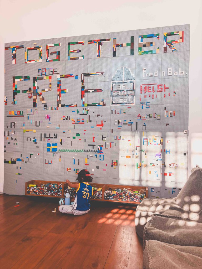 A child sits on a wooden floor at Family Nest Uluwatu, playing with LEGO bricks in front of a large wall filled with colorful LEGO creations and messages, like TOGETHER and FREE, as sunlight streams through a nearby window.
