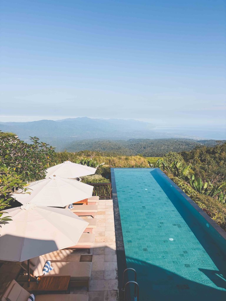 Infinity pool at Munduk Moding Plantation overlooks a lush green valley with mountains and blue sky in the distance. Several sun loungers with white umbrellas are set up beside the pool on a stone patio.