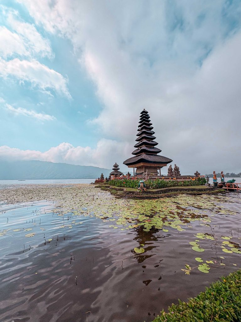 The traditional multi-tiered Ulun Danu Beratan temple stands on a small island in a serene lake, surrounded by water lilies, under a partly cloudy sky with mountains in the background.