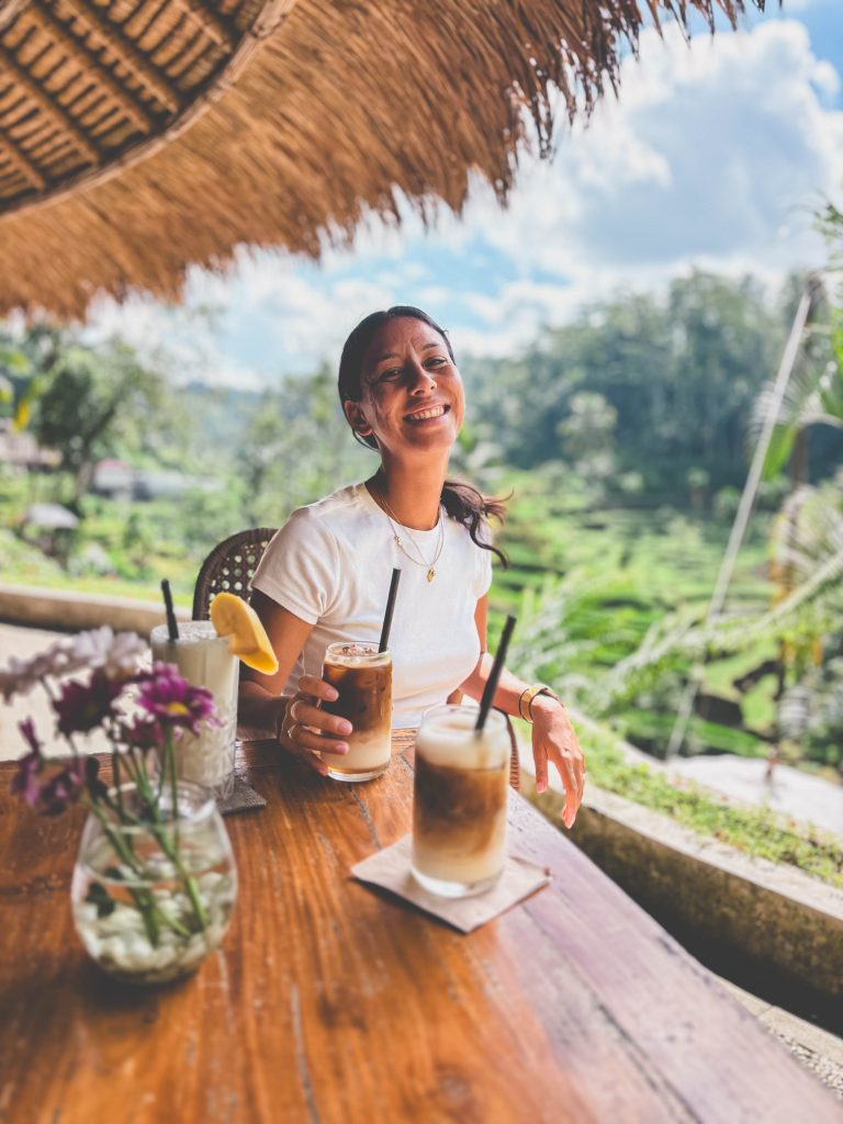 A woman in a white shirt smiles while sitting at a wooden table with iced drinks and flowers, outdoors at Tis Cafe Ubud with lush green scenery and rice terraces in the background.