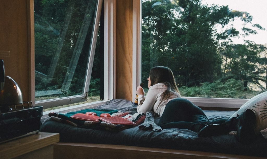 A woman lies on a cozy bed by a large open window, drinking from a mug and looking out at the lush green trees of Kangaroo Valley outside. A kettle and a tray with food are nearby.