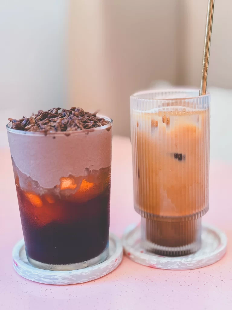 Two iced drinks on coasters: one is a dark beverage with frothy cream and chocolate shavings on top, the other is a lighter, creamy iced drink with a gold straw, both served on a pink surface—capturing the vibrant and inviting atmosphere of Kynd Cafe Uluwatu.