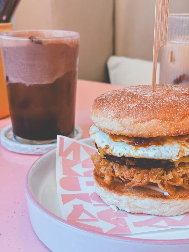 A breakfast sandwich with fried egg and crispy onions sits on a plate next to a glass of iced chocolate on a pink table at Kynd Cafe.