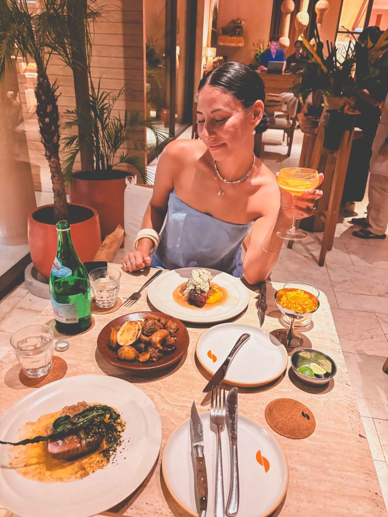 A woman in a blue top sits at a Teja Uluwatu Restaurant table with various dishes, holding up a cocktail and smiling. Potted plants and warm lighting decorate the background.