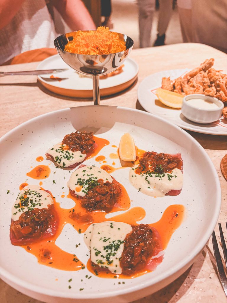 A plate with five ravioli topped with sauce and herbs, a wedge of lemon, a metal bowl of orange chips, and a side of fried food with dipping sauce and lemon slices—each thoughtfully plated in the signature style of Teja Uluwatu Restaurant.