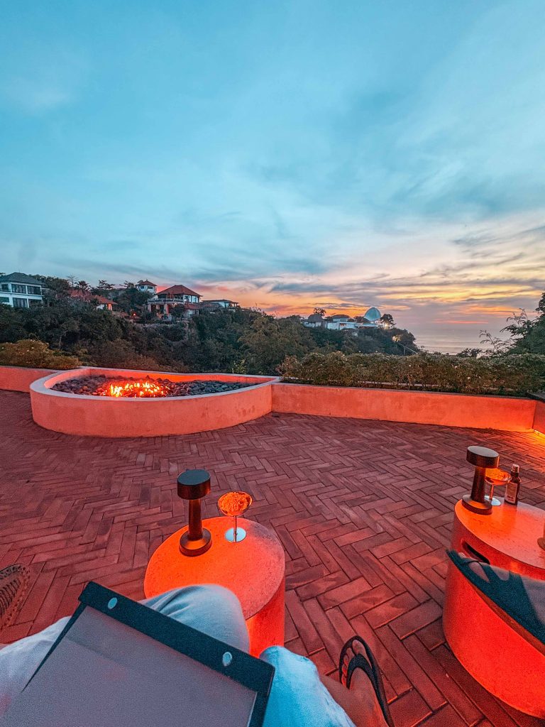 A person relaxes on a patio at sunset at Teja Uluwatu, with a fire pit, drinks on round tables, and a scenic view of hillside houses and the ocean in the distance under a colorful sky.