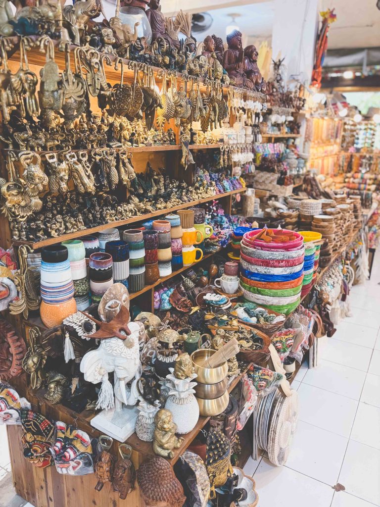 A market stall at the Uluwatu markets displays colorful baskets, mugs, brass and wooden figurines, and various handcrafted souvenirs arranged neatly on shelves and tables.