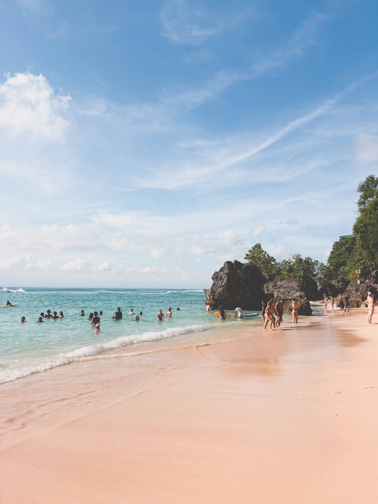 People enjoy swimming and relaxing on the sandy beach of Padang Padang Uluwatu, with clear turquoise water and rocky formations, under a sunny blue sky with a few clouds.