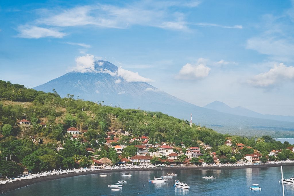 Amed’s coastal village features red-roofed houses and boats drifting in the water, all set against a lush green hillside and a large, cloud-topped volcano beneath a brilliant blue sky.