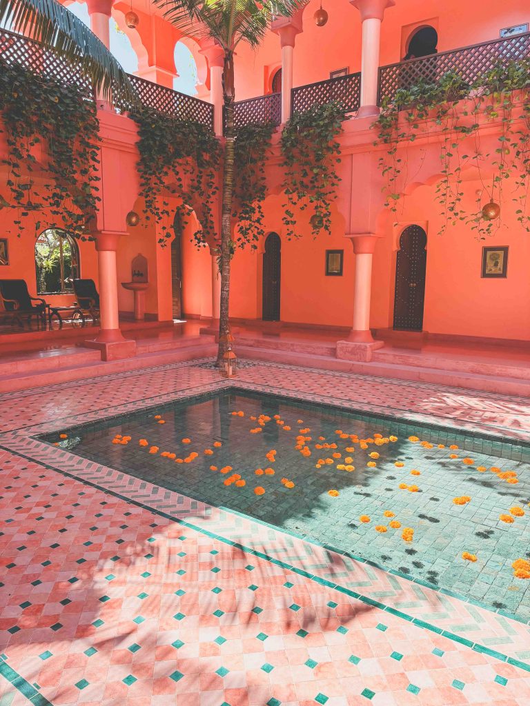 A courtyard with pink walls, green vines, arched doorways, and a small tiled pool reminiscent of the tranquil ambiance at Bodyworks Spa Seminyak. Yellow and orange flowers float on the water as sunlight streams in, casting soft shadows throughout the peaceful oasis.