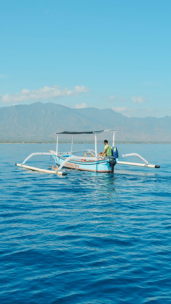 A small outrigger boat with a canopy floats on calm blue water, with a person onboard and mountains visible in the background under a clear sky—a serene scene often found near Lovina, one of the best areas to stay in Bali.