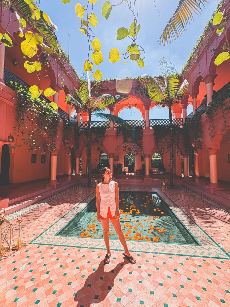 A person in a white top and red skirt stands by a pool with floating flowers in a sunlit courtyard at Seminyak Bodyworks, surrounded by pink walls, arches, greenery, and palm trees.