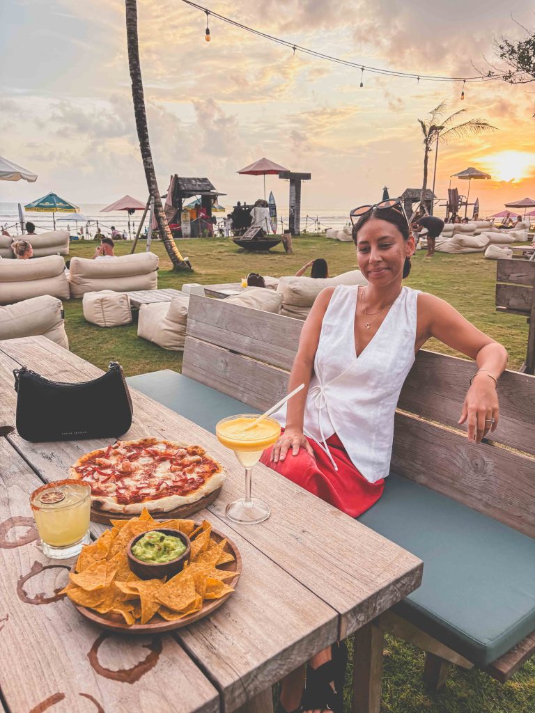 A woman sits at an outdoor beachside table at sunset, with pizza, nachos and guacamole, and two cocktails in front of her. She is relaxed, wearing a white blouse and red skirt. The atmosphere is casual and inviting—perfect for unwinding after exploring all the things to do in Seminyak or savoring the ambiance of a sunset beach club.