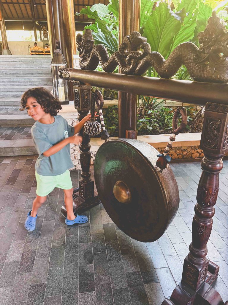 A young child in a blue shirt and light shorts is striking a large bronze gong with a mallet in an ornate, tropical setting with green plants and wooden pillars—one of the best things to do in Bali with kids at the Hyatt Regency Sanur.